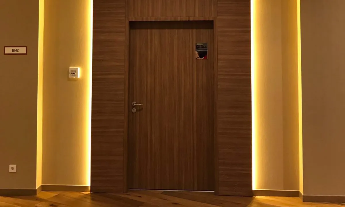 Puerta de madera oscura con su marco revestido hasta el techo, entre dos tiras de luz cálida, en el NH Collection Berlin Mitte