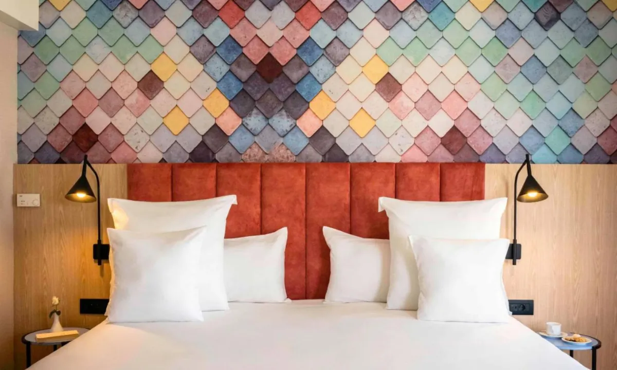 Cama del Barceló Fez Medina con cabecero tapizado en terracota bajo una pared de escamas de colores, entre paneles de madera clara y dos lámparas de brazo