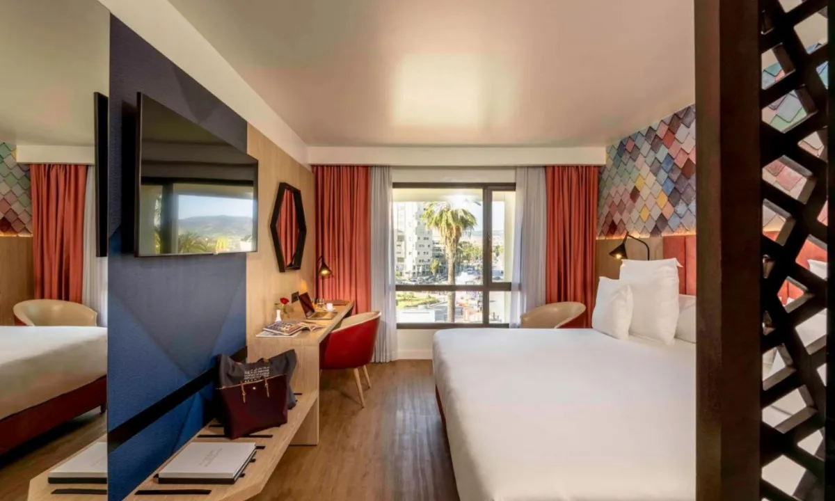 Habitación del Barceló Fez Medina con escritorio y estante de madera clara, cortinas rojas y una ventana con palmeras al fondo