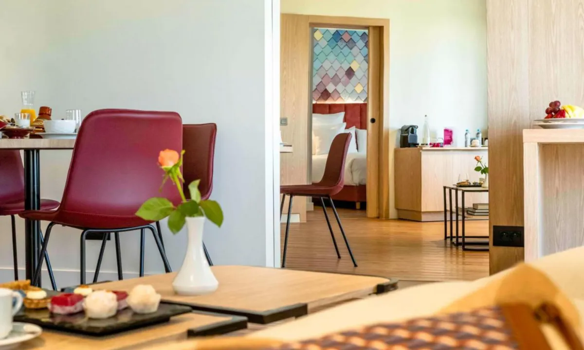 Zona de estar del Barceló Fez Medina con mesa de desayuno y sillas granate; al fondo, por la puerta corredera, el dormitorio con la pared de escamas de colores