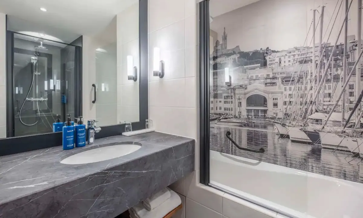 Baño del Radisson Blu Marsella con lavabo sobre encimera gris, bañera y un fotomural en blanco y negro del puerto