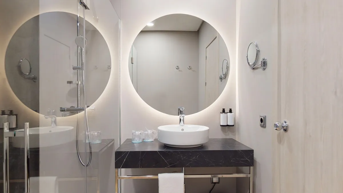 Baño del Occidental Sevilla con espejo redondo iluminado, lavabo sobre encimera oscura y ducha